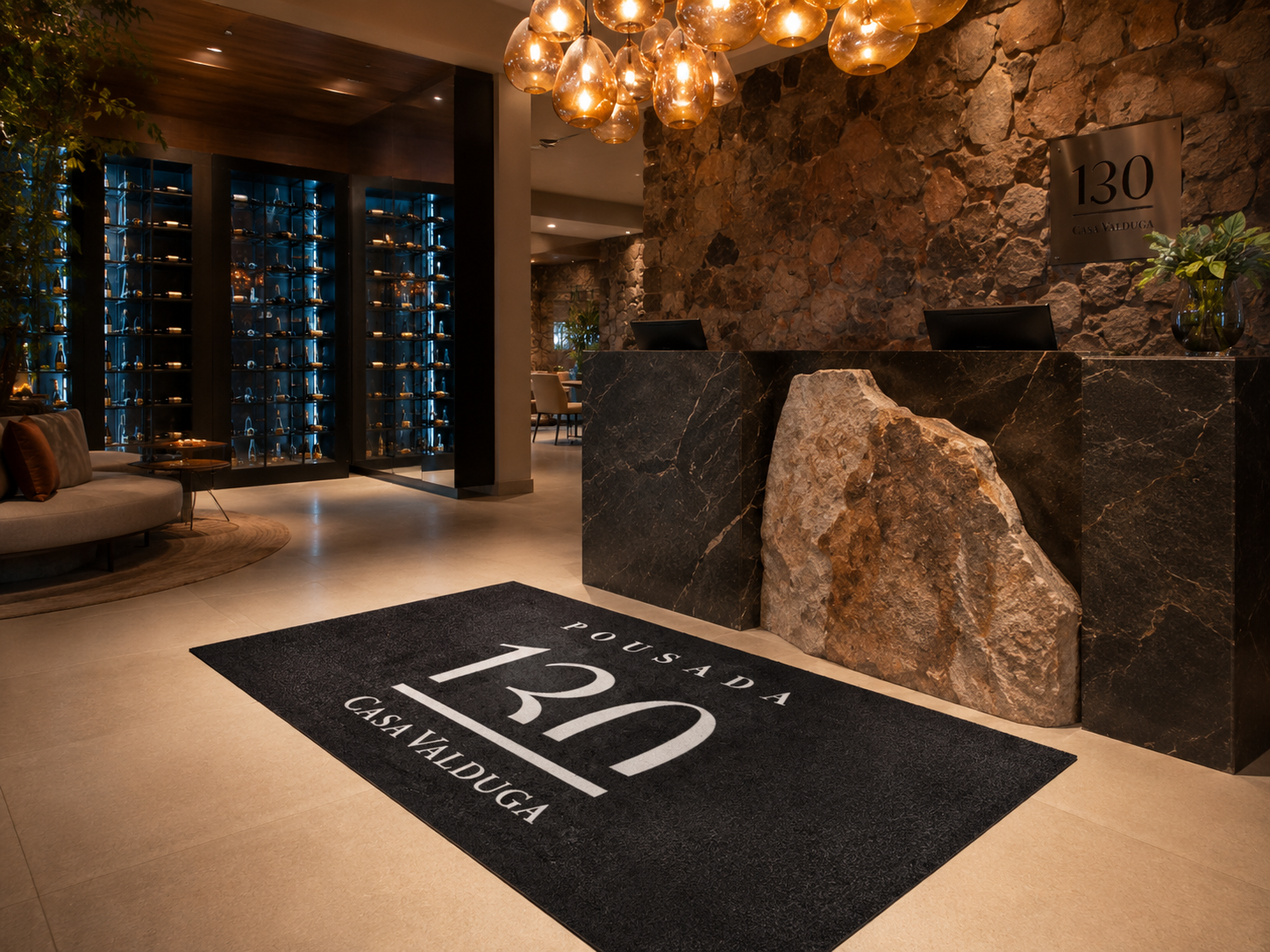 Capacho emborrachado preto personalizado com o texto Pousada 130 Casa Valduga em branco, posicionado no piso de recepção sofisticada com parede de pedra e iluminação quente, fotografia realista de ambiente hoteleiro de alto padrão