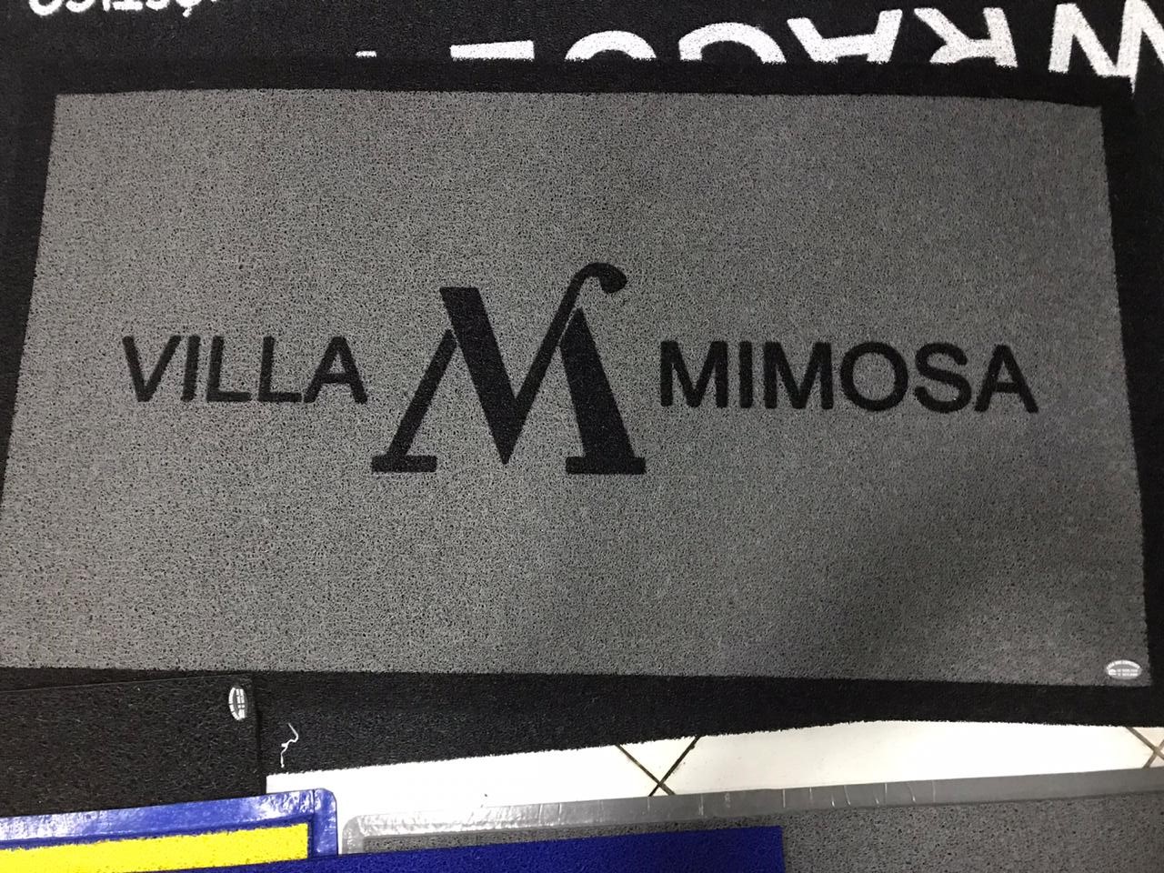 Tapete de entrada em vinil cinza personalizado com o texto Villa Mimosa em preto e detalhe de monograma, textura lisa e fácil de limpar, fotografia realista em ambiente de recepção