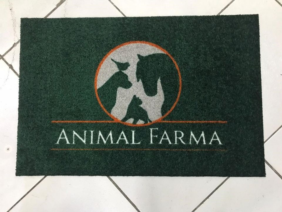 Capacho personalizado verde escuro com a logomarca Animal Farma, círculo prateado com silhuetas de animais em contorno laranja, sobre piso de cerâmica clara, fotografia realista de tapete para clínica veterinária de linha profissional