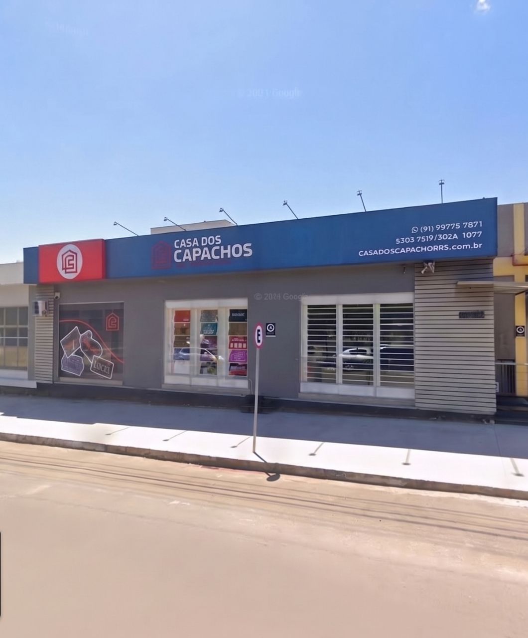 Fachada da loja Casa dos Capachos com letreiro azul destacando o nome da empresa, logomarca vermelha e branca em painel lateral, vitrines com produtos expostos, calçada e rua em dia ensolarado de céu azul claro, fotografia realista de fachada comercial