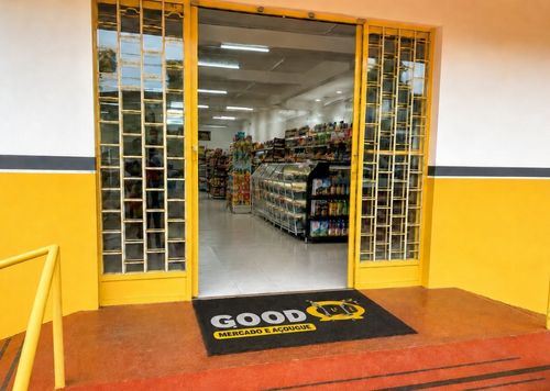 Capacho de fibra preto e amarelo com a logomarca Good Mercado e Açougue, instalado na entrada de mercado com portas de vidro amarelas e prateleiras ao fundo, fotografia realista de fachada comercial de alta circulação